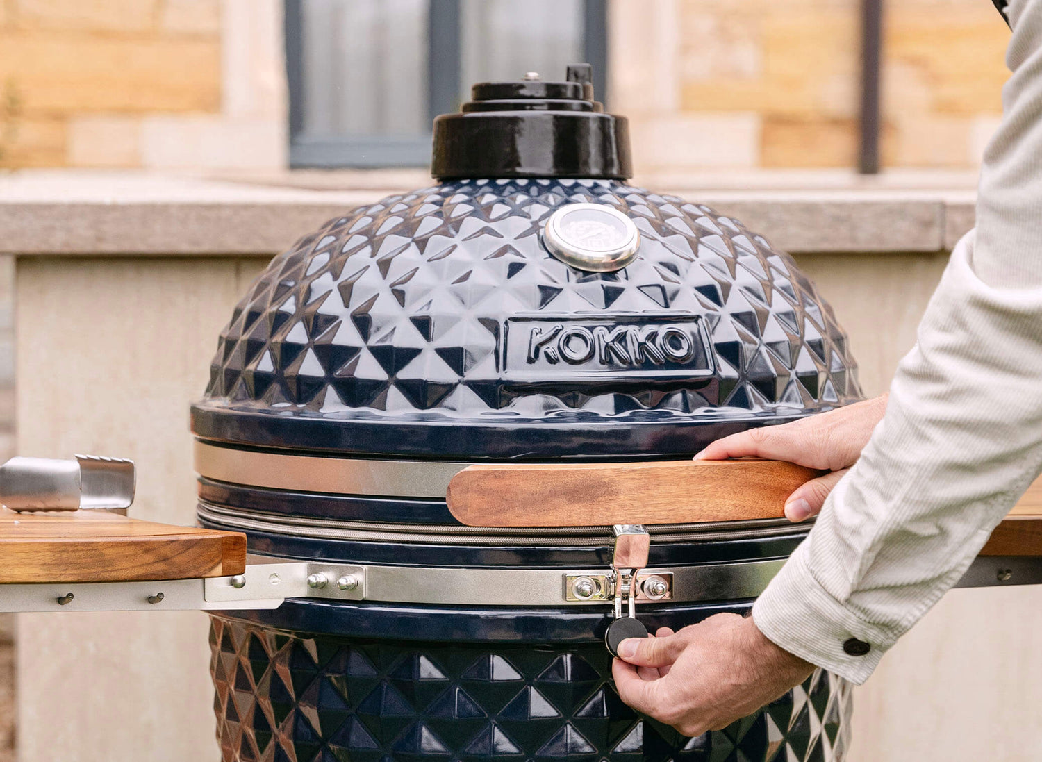 Hand die het veiligheidsslot van een zwarte Kokko kamado bedient, met een deksel van getextureerd keramiek, houten handgreep en geïntegreerde thermometer, in gebruik buitenshuis.