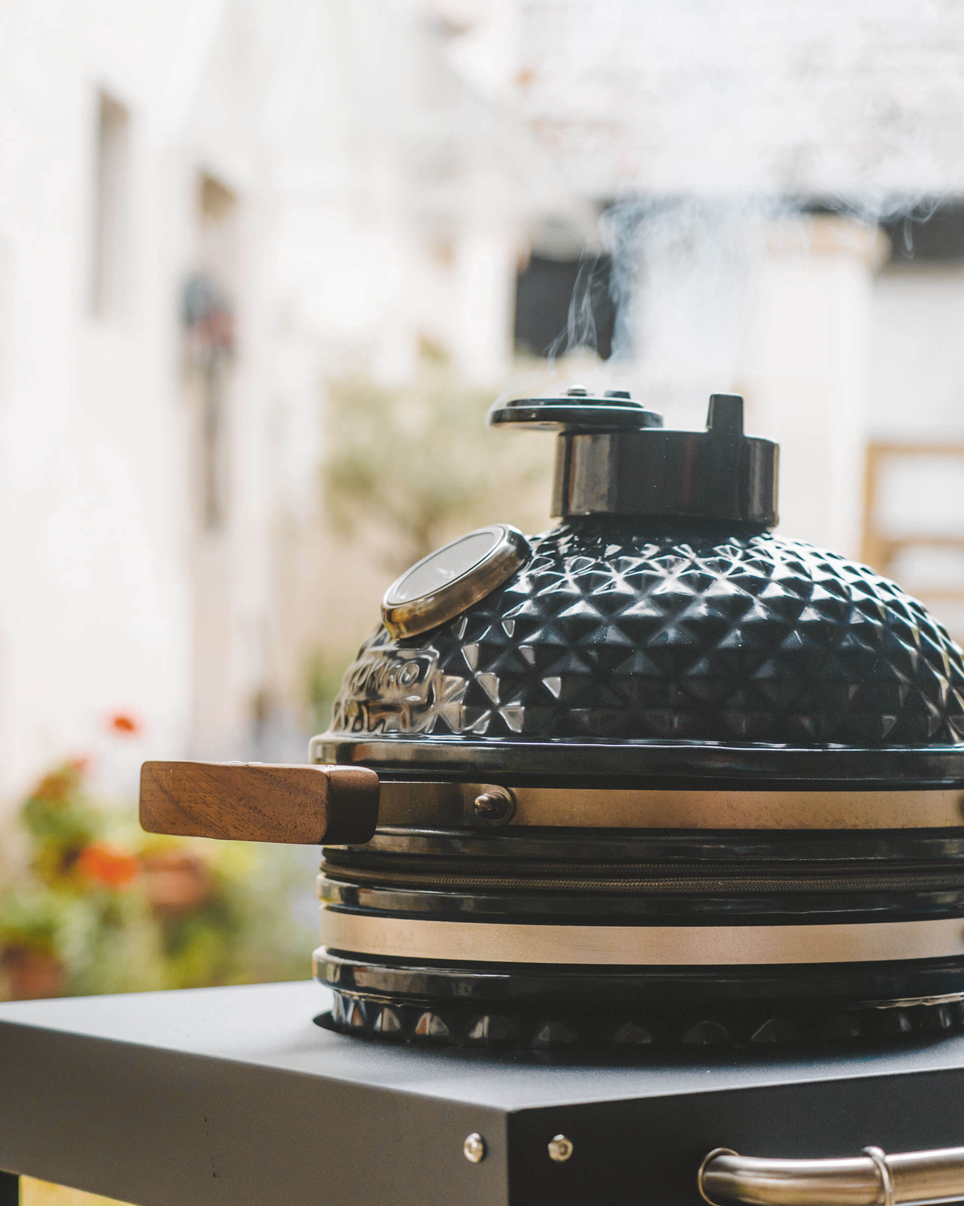 Zwarte Kamado Kokko op een onderstel, met een deksel van getextureerd keramiek, houten handgreep, geïntegreerde thermometer en rook die uit de schoorsteen komt.