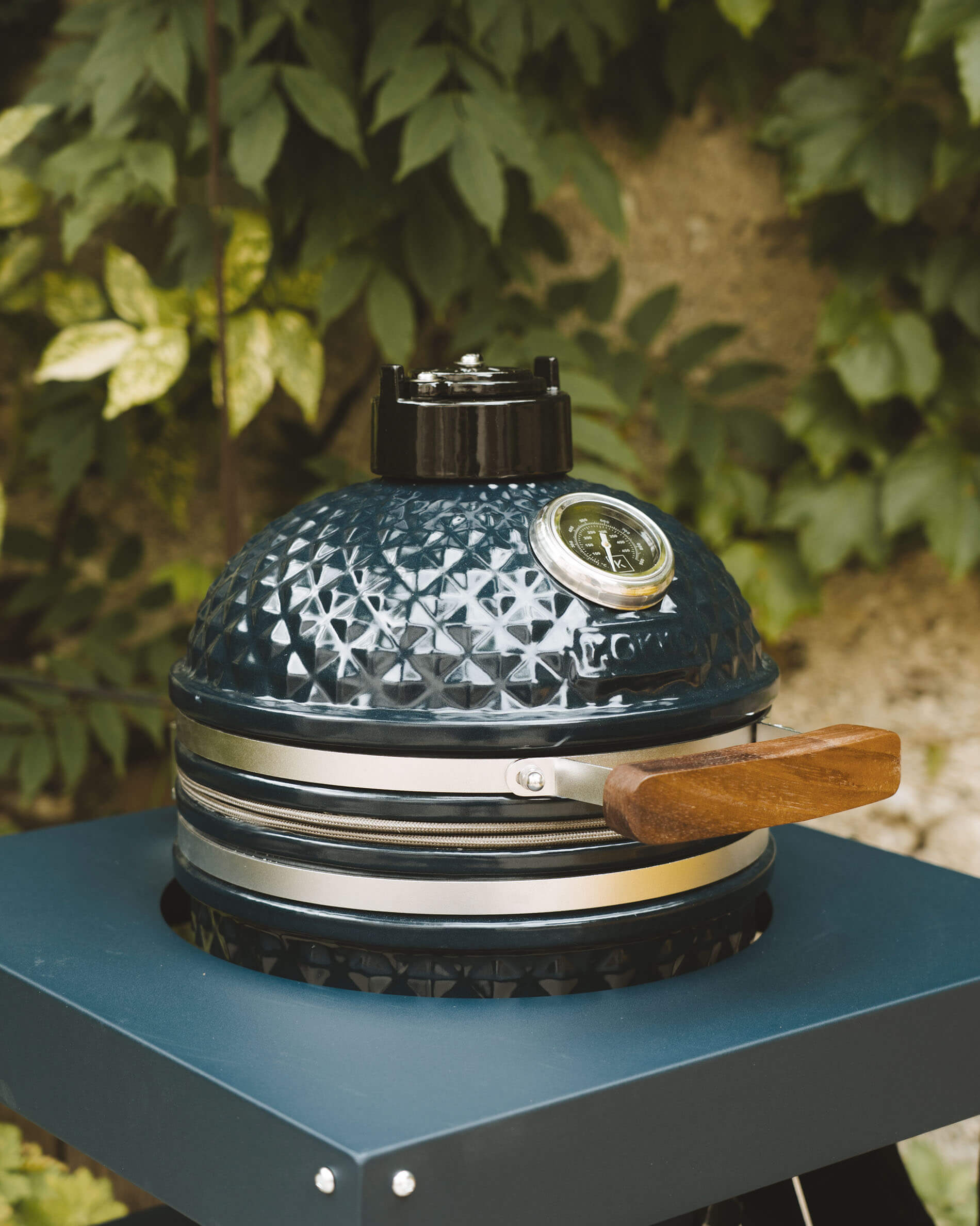 Vooraanzicht van een Kokko kamado in oceaankleur op een onderstel, met de nadruk op het getextureerde keramiek, de geïntegreerde thermometer en de houten handgreep.