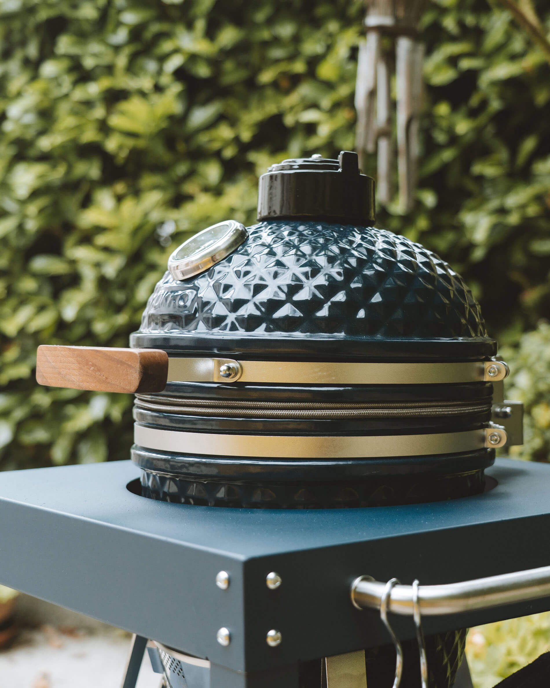 Zijaanzicht van een Kokko kamado in oceaankleur op een verrijdbaar onderstel, met de keramische deksel, houten handgreep en geïntegreerde thermometer in de kijker.
