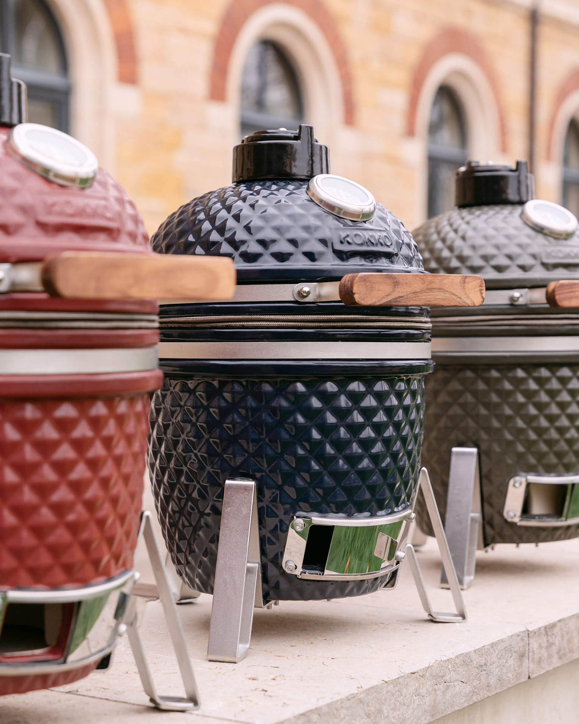 Kamado’s van Kokko in gestructureerd keramiek, gepresenteerd in diverse kleuren met houten handgrepen en metalen onderstellen.